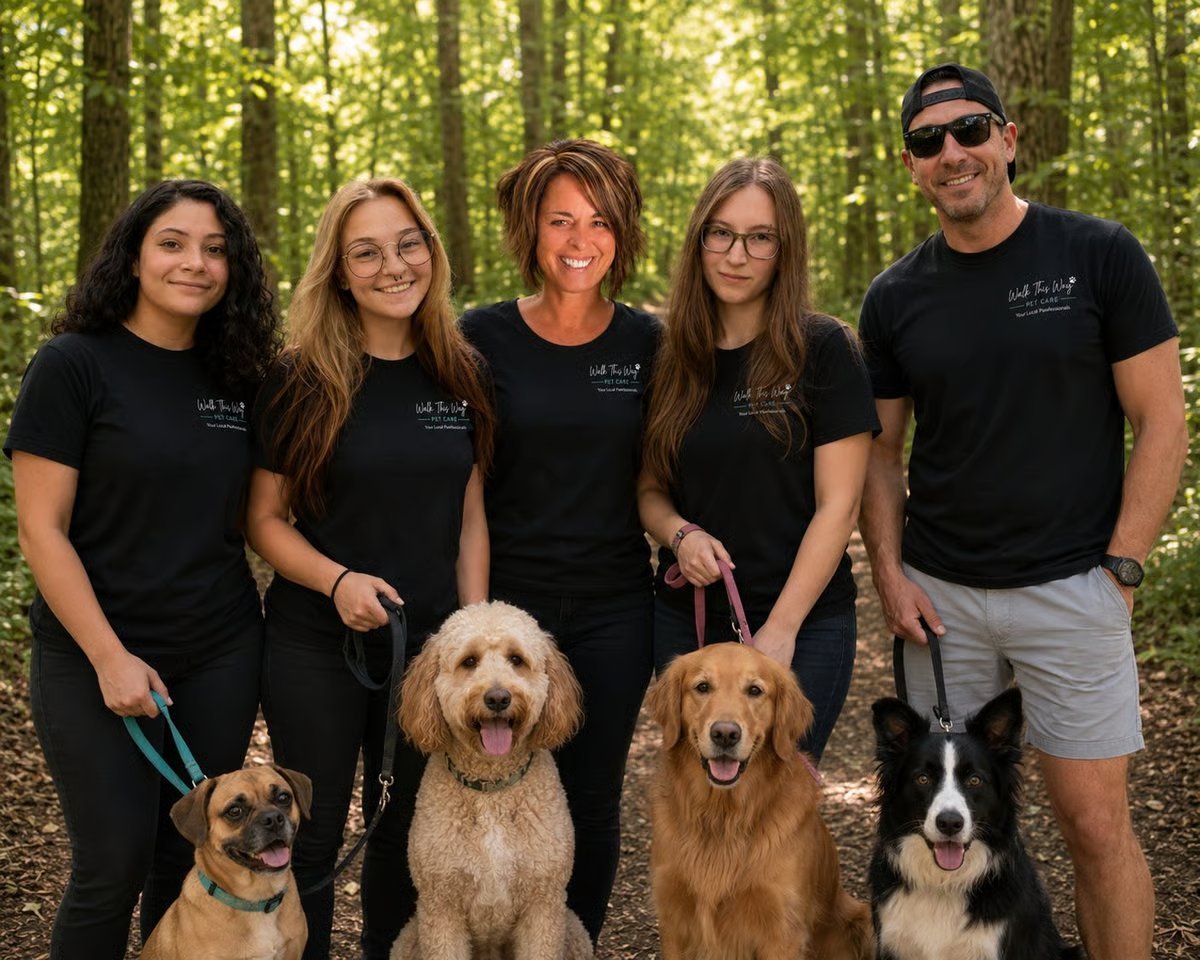 Five people in matching black t-shirts pose with four dogs on a forest trail lined with tall trees